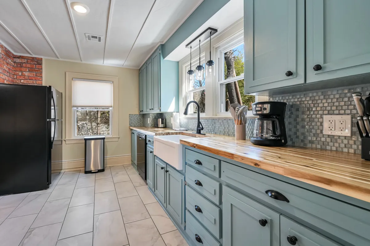This image features the kitchen's blue cabinetry, a large farmhouse sink, wooden countertop, and modern black appliances. The exposed brick accent wall adds character, and the overall space is warm and welcoming, inviting people to gather for meal preparation and conversation.