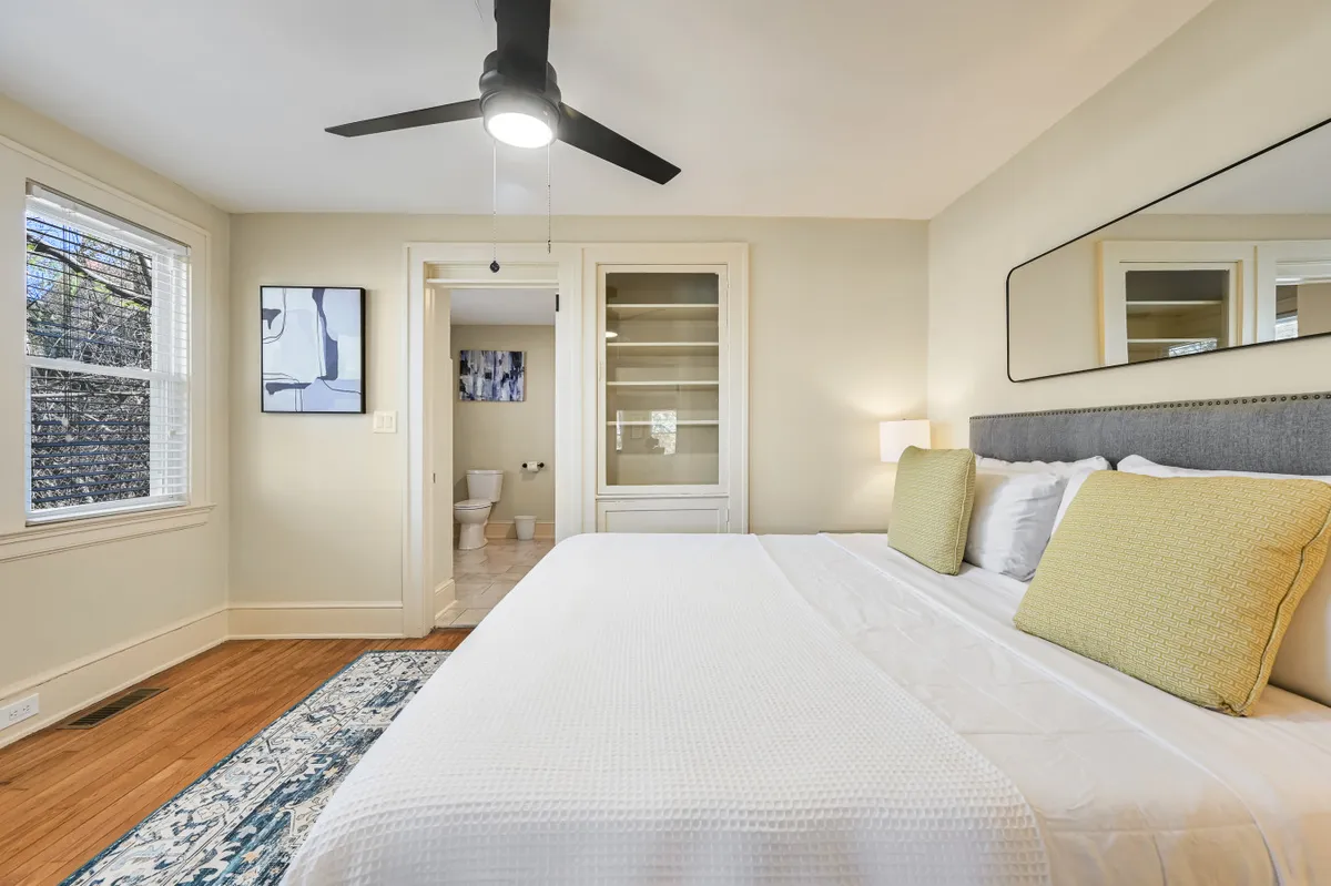 A cozy bedroom featuring a bed with yellow accent pillows, a ceiling fan, framed artwork, a large mirror, and wooden flooring. The space is bathed in natural light, creating a warm and inviting atmosphere for reading or unwinding.