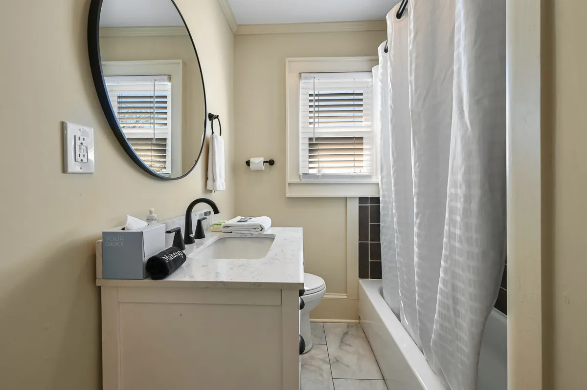 A calming bathroom scene with a round mirror, vanity with sink, shower curtain, and a window with blinds. The clean, serene setting offers ample space for personal routines and relaxation, echoing the stress-free, modern amenities available to groups.