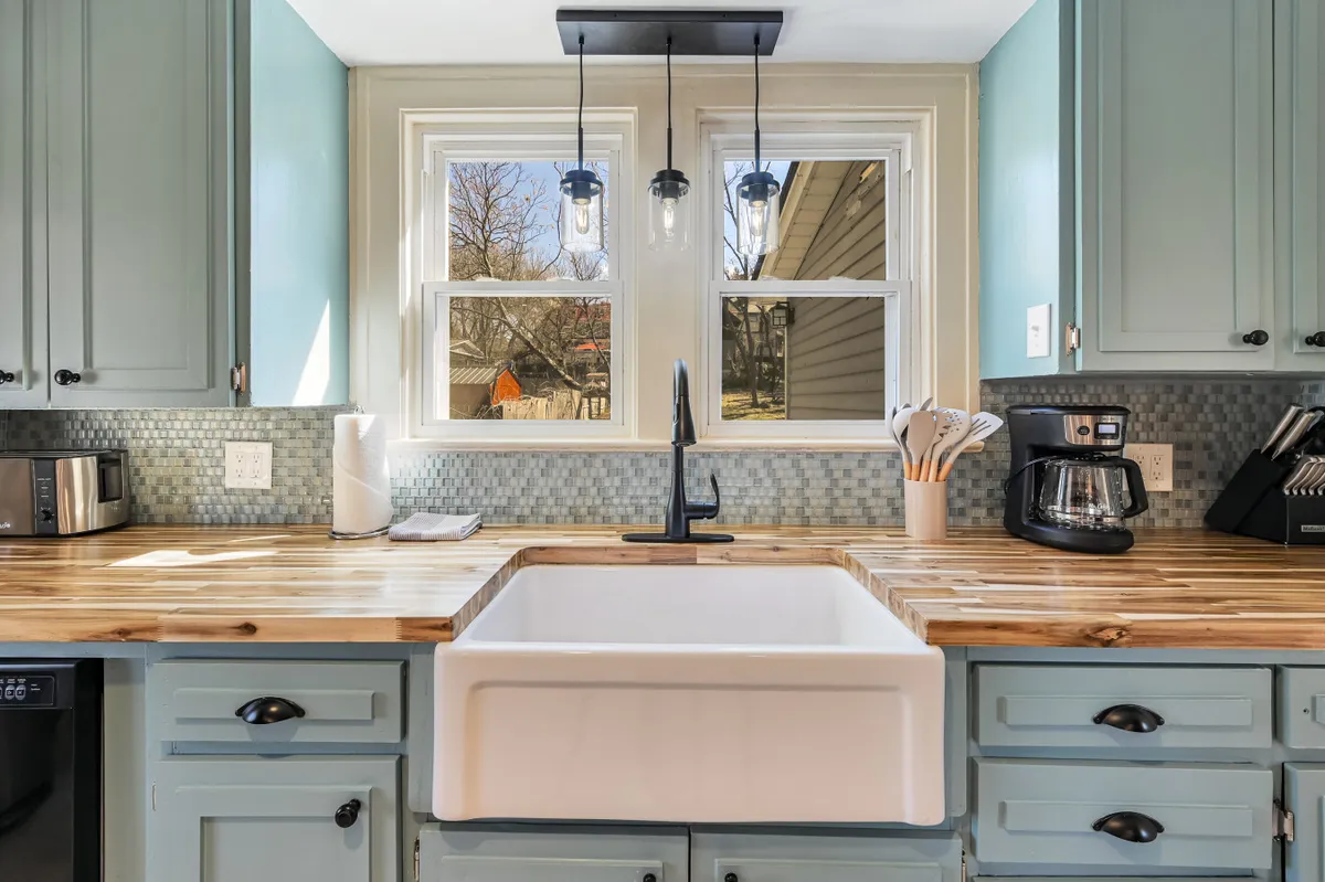 The kitchen scene displays a farmhouse sink, wooden countertops, modern kitchen appliances, pendant lighting, and window views, all in a cozy, inviting atmosphere. The setup is ideal for group cooking, baking, or enjoying coffee together.