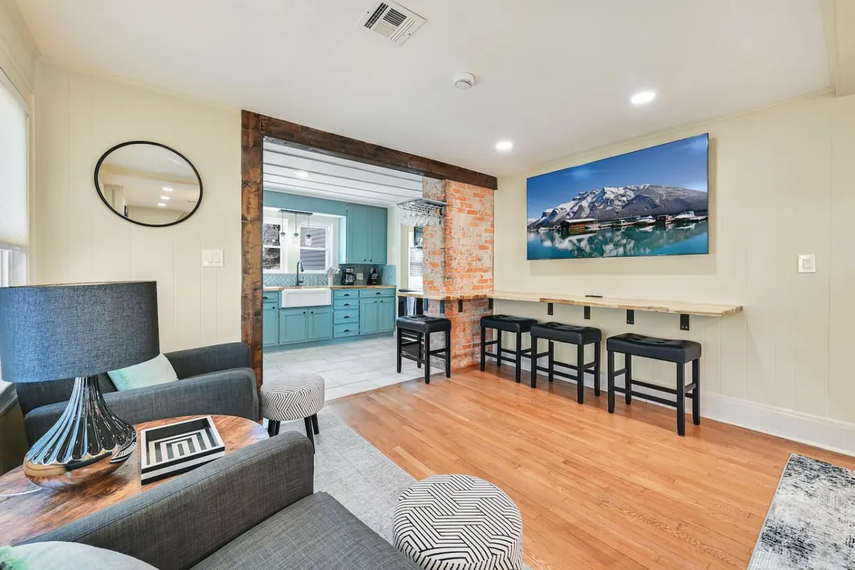 A spacious, light-filled living area featuring a comfortable sofa with a striped cushion, a modern black lamp, and a wooden bar seating area adjacent to a kitchen with blue cabinets. A large mountain landscape artwork provides a nod to the surrounding environment. The setting is inviting and ideal for socializing, entertaining, or relaxing, with multiple seating arrangements that encourage group interaction.
