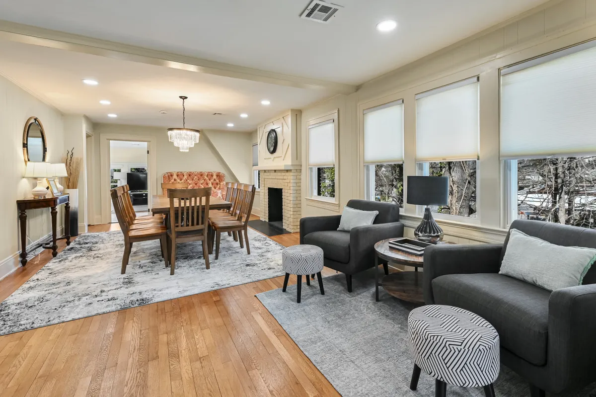 A wide, welcoming view of an open-plan space featuring a dining table beneath a chandelier, surrounded by a comfortable sofa set, a cozy fireplace, and a decorative rug. The seamless transition between living and dining areas is evident, and the décor blends historic charm with modern furnishings.