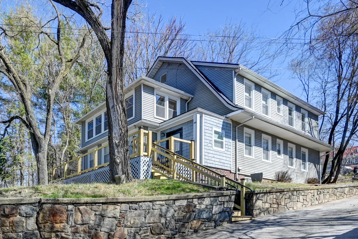 An inviting view of the home's exterior, highlighting a stone wall, wooden steps, large windows, and a canopy of trees. The image conveys the sense of a welcoming entrance and the beauty of the surrounding natural environment.