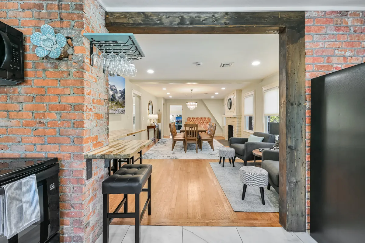 The image showcases a beautifully renovated interior featuring an exposed brick wall, a sleek wooden bar with stools perfect for entertaining, a spacious dining area for family dinners, and a cozy seating area inviting guests to relax. The scene exudes a warm and welcoming ambiance, ideal for gatherings and celebrations.