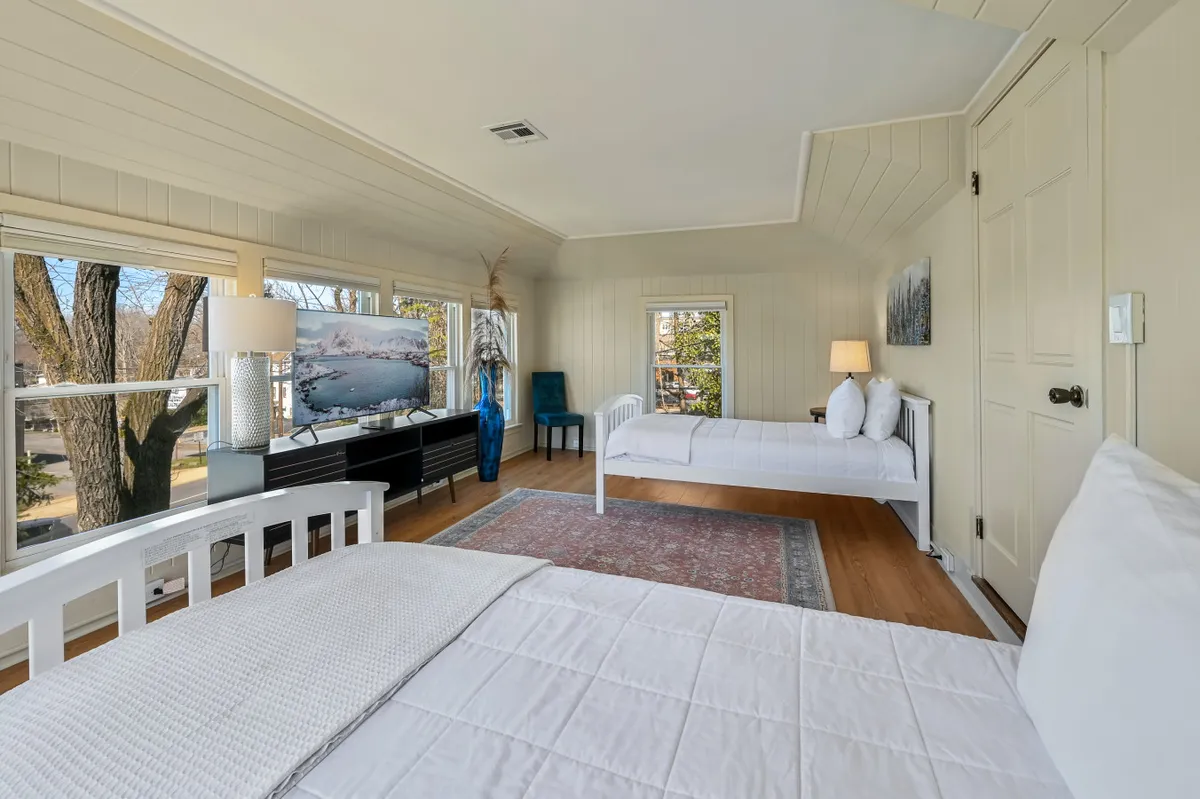 A serene bedroom with large windows framing scenic views, a pair of white beds, a decorative lamp, and a TV for modern comfort. A blue vase with plants adds a touch of freshness. The space feels airy and tranquil, ideal for quiet reflection, reading, or enjoying a relaxing evening with entertainment.