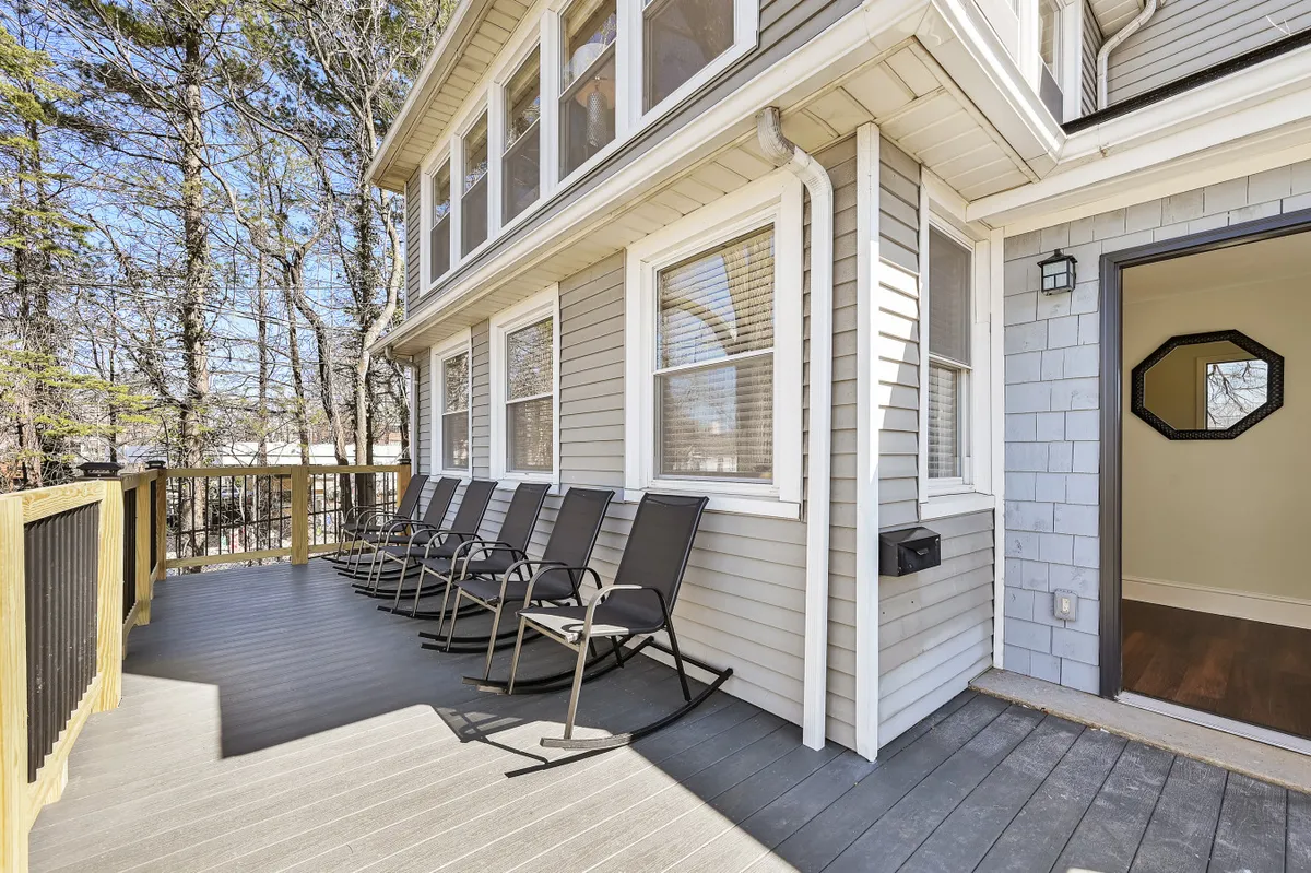 A row of classic rocking chairs lined up along a deck, with the tranquil backdrop of tall trees and sunlight filtering through. The setting exudes calm and invites guests to relax and enjoy the natural surroundings.