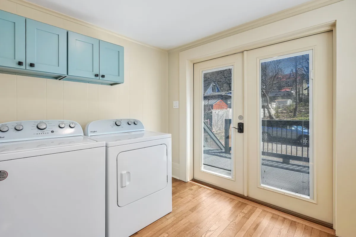 A bright, functional utility area with a washer and dryer set, blue cabinetry, and double doors that open directly to the outdoors, inviting natural light. The open doors create a seamless transition between indoor and outdoor spaces, hinting at opportunities for fresh-air breaks or easy access for off-site inspiration walks.