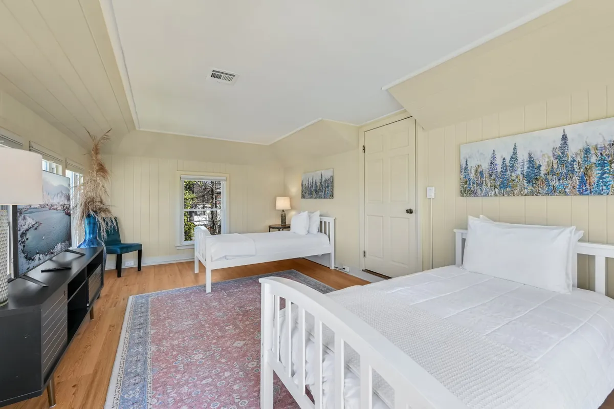 A bright, inviting bedroom featuring two white beds with a decorative rug, lamp, and tasteful artwork on the wall. Natural light pours in, highlighting the classic molding and hardwood flooring that evoke the home's historic character. The arrangement is welcoming for families or friends sharing a room, while also feeling peaceful and private.